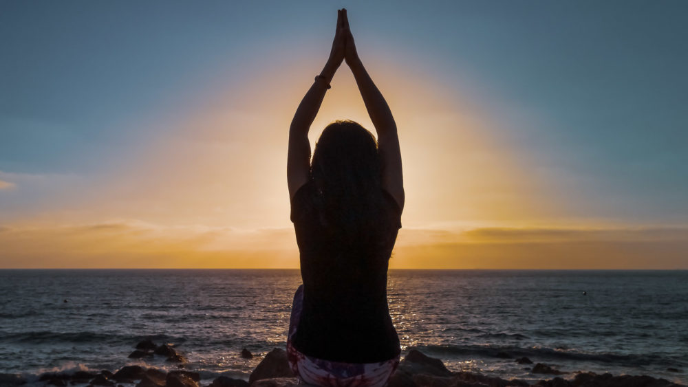 Une femme pratiquant la posture du lotus du Hatha yoga au coucher du soleil devant la plage